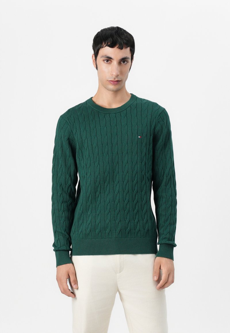 Zeleni pulover z pletenim vzorcem in okroglim ovratnikom, z rebrastimi manšetami in robom, ki ima majhen logotip na prsih. Model nosi svetle hlače.