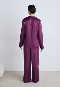 Violet satino loungewear komplektas, kuriame yra ilgomis rankovėmis marškiniai su subtiliu raštu ir plačios kelnės, parodytas iš nugaros prieš neutralų foną.