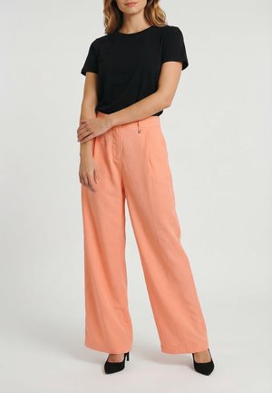 Femme portant un haut noir à manches courtes associé à un pantalon taille haute ample couleur pêche et des chaussures à talons noires, debout les bras croisés.