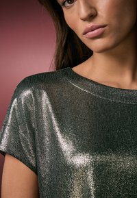 Skinande mörkgrön topp med ett mjukt, texturerat tyg och en vid halsringning. Kortärmad med reflekterande detaljer som ger ett lager-på-lager-uttryck.