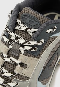 Γκρι και μπεζ αθλητικό παπούτσι με κορδόνια σε ασπρόμαυρο σχέδιο, ύφασμα από δίχτυ και μαύρο οβάλ λογότυπο στη γλώσσα.