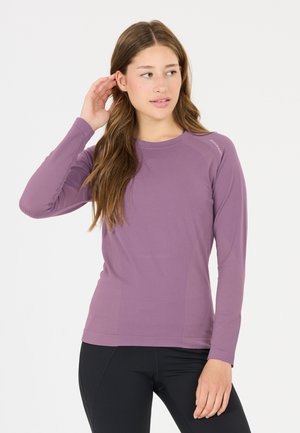Jonge vrouw met lang bruin haar, een paarse longsleeve en zwarte broek, raakt haar oor aan en kijkt opzij.