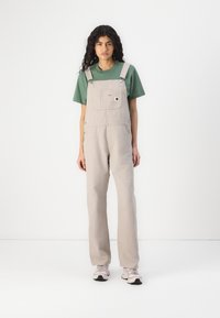 Lichtbeige overalls gemaakt van duurzaam materiaal, met een voorknop, verstelbare bandjes en een relaxte pasvorm. Gecombineerd met een groene T-shirt en roze sneakers.