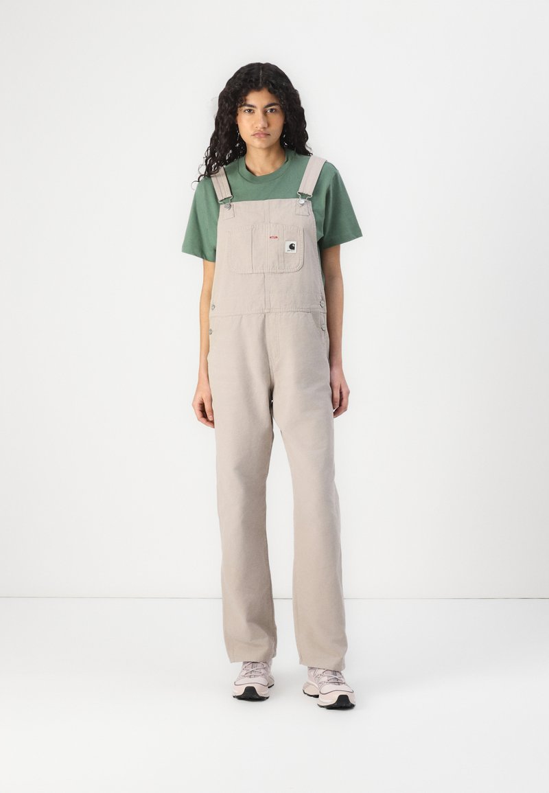 Lichtbeige overalls gemaakt van duurzaam materiaal, met een voorknop, verstelbare bandjes en een relaxte pasvorm. Gecombineerd met een groene T-shirt en roze sneakers.