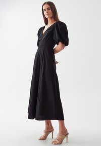 Gaun hitam panjang hingga lantai dengan lengan puff, leher V, dan bodice yang pas. Fabrik halus dengan sedikit kilau, dipadukan dengan sandal hak tinggi berwarna beige.