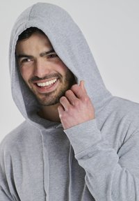 Grå hættetrøje lavet af struktureret stof, med en frontlomme og snoretræk. Modellen smiler og holder hætten.