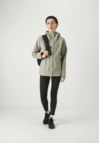 Casaco impermeável cinza claro com capuz, fecho de correr à frente e punhos ajustáveis; combinado com leggings pretas e ténis pretos.