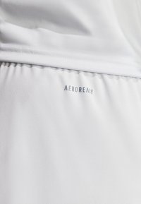 Vitt atletiskt tyg med en slät textur. Har "AEROREADY"-logotyp i ljusgrått vid fållen. Enkel, funktionell design.