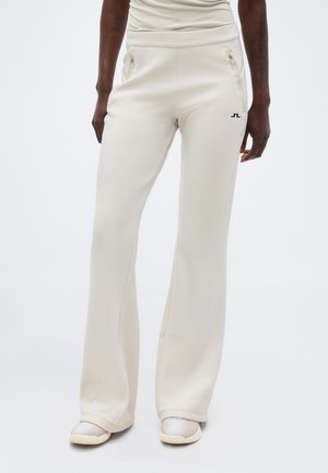 Άτομο που φορά κρεμ παντελόνια με φαρδιά φάρδια και τσέπες με φερμουάρ, καθώς και casual παπούτσια, στέκεται μπροστά σε απλό λευκό φόντο.