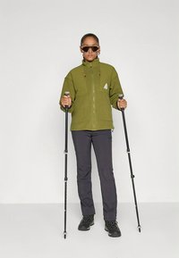 Flis jakna olivno zelene barve z zadrgo spredaj, dvema žepoma z zadrgo in logotipom; kombinirana s temnimi hlačami in črnimi pohodnimi čevlji, v roki drži pohodne palice.