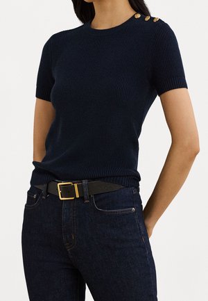 Pull en tricot côtelé bleu marine à manches courtes avec des boutons dorés sur l'épaule, associé à un jean foncé et une ceinture noire avec une boucle en or.