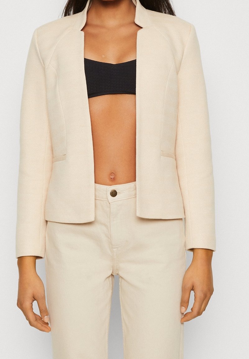 Blazer de color beige claro con un patrón de espiga, corte ajustado y sin botones. Se lleva sobre un bralette negro y pantalones de color claro.