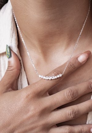 Silberne Halskette mit einer geschwungenen Kette, besetzt mit kleinen, klaren Steinen; auf der Haut getragen, von einer Hand mit texturierter Haut und lackierten Nägeln gehalten.