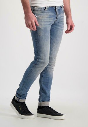 Blauwe denim jeans met een slim fit, gekenmerkt door lichte slijtage, een typische vijf-pocket ontwerp en versleten uiteinden. Gepaard met zwarte sneakers.