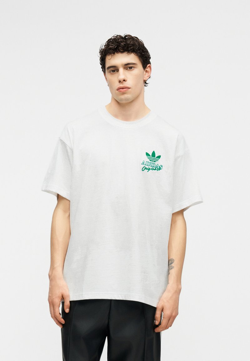 Jeune homme aux cheveux bouclés portant un t-shirt blanc Adidas Originals avec un logo vert et un pantalon noir, debout devant un fond uni.
