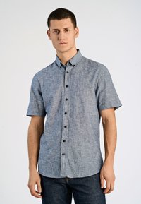 Camicia a maniche corte con bottoni, realizzata in tessuto chambray blu con una texture sottile; presenta bottoni marroni e un colletto a punta.