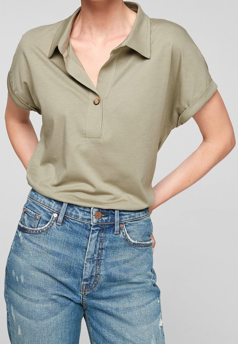 Personne portant un polo à manches courtes couleur olive clair avec un bouton et des manches retroussées, associé à un jean bleu déchiré taille haute.
