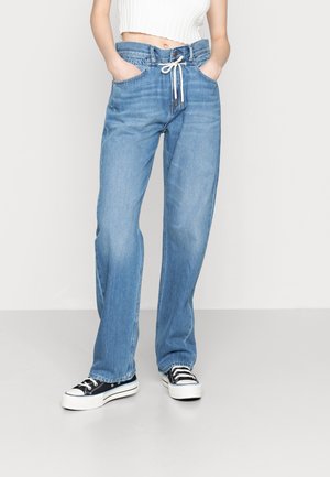 Persoon die lichtblauwe high-waisted wijde jeans draagt met een witte trekkoord, wit cropped topje en zwarte lage sneakers.