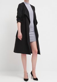 Czarny płaszcz trench na dopasowanej, jasnoszarej sukience o teksturowanym wzorze. Płaszcz ma luźny krój z otwartym przodem i długimi rękawami.