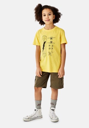 Gul bomulds-t-shirt med skateboardgrafik, båret med olivengrønne shorts. Modellen har også hvide sneakers og grå sokker med rød tekst.