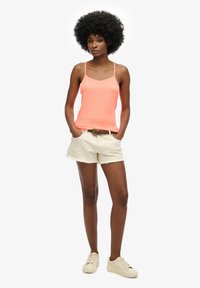 Camisola arancione neon con spalline sottili, abbinata a pantaloni corti crema con orli sfrangiati e sneakers chiare. Design semplice e casual.