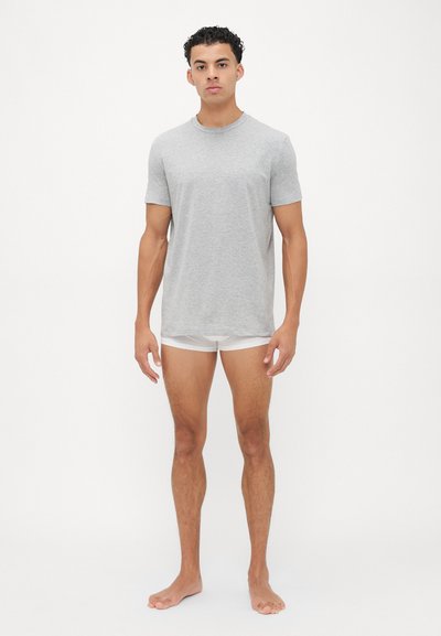 Graues Baumwoll-T-Shirt mit kurzen Ärmeln und Rundhalsausschnitt, getragen mit weißen Shorts, präsentiert an einer Person, die barfuß vor einem weißen Hintergrund steht.