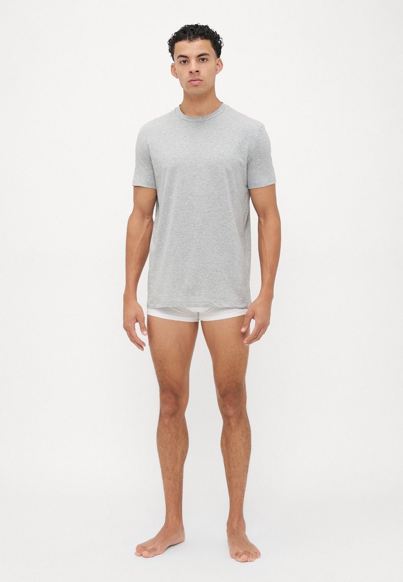 Graues Baumwoll-T-Shirt mit kurzen Ärmeln und Rundhalsausschnitt, getragen mit weißen Shorts, präsentiert an einer Person, die barfuß vor einem weißen Hintergrund steht.