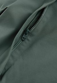 Țesătură verde închis cu o textură netedă, având un fermoar contrastant cu accente negre, parțial deschis pentru a dezvălui interiorul.