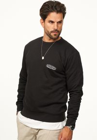 Black sweatshirt with round neckline, long sleeves, and ribbed cuffs. Features a white embroidered logo on the left chest. Worn over a white shirt.