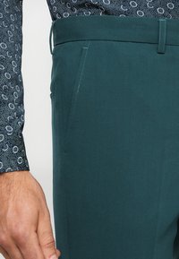 Tirkizne pamučne hlače s bočnim džepovima, s glatkom teksturom i krojem po mjeri, u kombinaciji s košuljom dugih rukava s uzorkom.
