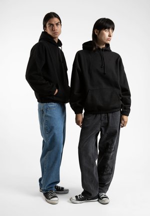 Two individuals wear oversized black hoodies. One pairs a black hoodie with dark jeans featuring a subtle pattern, the other wears light blue jeans.