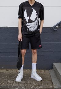Black mesh jersey with white graphic design, paired with black shorts. White high-top sneakers and black sling bag complete the outfit.