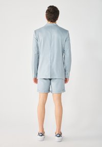 Blazer azzurro chiaro e pantaloni corti abbinati, vestibilità su misura, texture morbida, spacco singolo, maniche arrotolate, indossato con sneakers bianche.