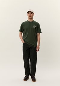 Zelené bavlněné tričko s bílým logem na hrudi, doplněné tmavými džínami a hnědými botami. Béžová kšiltovka nošená nakloněná vpřed.