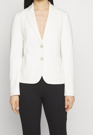 Žena oblečená v přiléhavém bílém blejzru se dvěma knoflíky a předními kapsami, v kombinaci s černými kalhotami, stojící před jednobarevným pozadím.