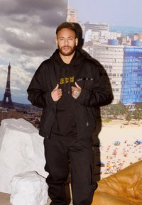Giacca nera oversize sopra un felpa nera con scritte gialle. Pantaloni scuri con tasche, in piedi di fronte a uno sfondo panoramico.