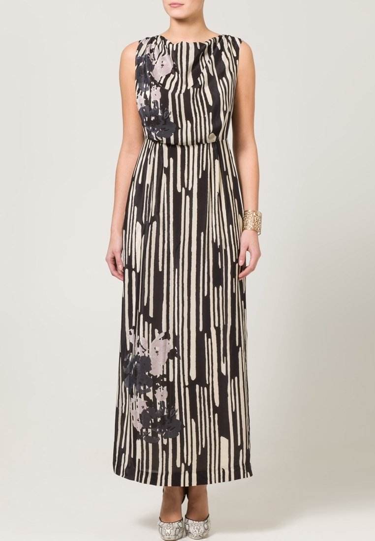 Femme portant une robe longue sans manches à rayures noires et beiges avec des accents floraux et un bracelet manchette en or à son poignet droit.