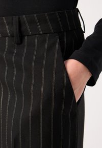 Mustat räätälöidyt housut, joissa on pinstripe-kuvio, vyö ja taskut. Kankaan pinnassa on tekstuuri ja kontrastiväriset valkoiset ompeleet.