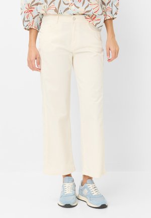 Kvinde iført off-white bukser med lige ben, en blomstret top og lyseblå sneakers, der står på en ensfarvet baggrund.