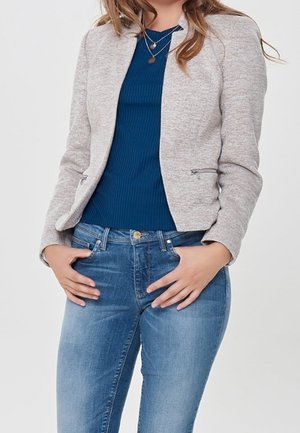 Femme portant un blazer gris clair zippé, un haut bleu côtelé, un jean bleu et des colliers en or superposés, les mains sur les hanches.