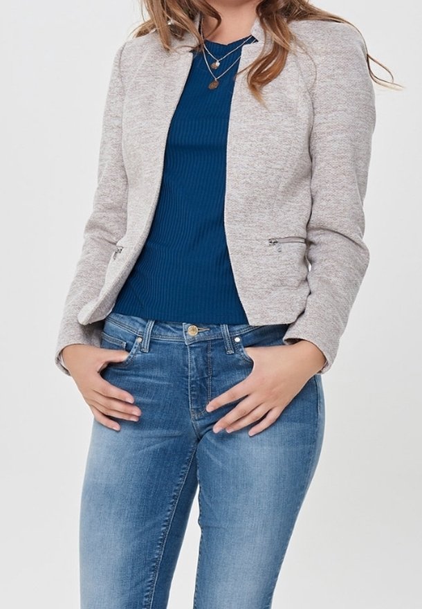 Femme portant un blazer gris clair zippé, un haut bleu côtelé, un jean bleu et des colliers en or superposés, les mains sur les hanches.
