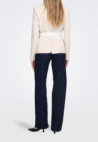 Femme aux longs cheveux blonds portant un blazer beige avec une ceinture en dentelle blanche, un jean large foncé et des chaussures noires à petits talons, vue de dos.