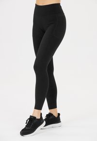 Vrouw in zwarte hoge tailleleggings met subtiel patroon en zwarte hardloopschoenen met witte zolen tegen een witte achtergrond.