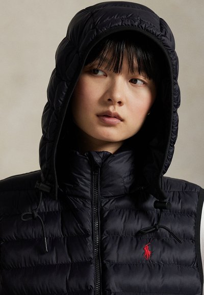 Modèle portant une doudoune noire matelassée avec une grande capuche, des cordons de serrage et un logo Polo brodé en rouge.