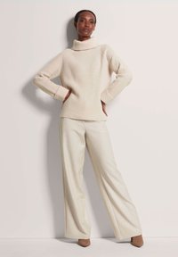Beige Ripp-Turtleneck-Pullover mit weiten Ärmeln, kombiniert mit hellbeigen hochgeschnittenen weiten Hosen und spitzen Absatzschuhen.