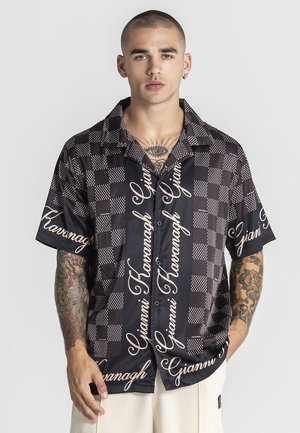 Mladý muž s oholenou hlavou a tetovaniami má na sebe čiernobielu košeľu s krátkym rukávom a vzorom kociek so skriptovým textom, stojaci na jednoduchom pozadí.