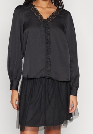 Blouse noire à manches longues, avec des détails en dentelle le long de l'encolure, et une superposition de jupe en tulle. Tissu lisse avec une coupe décontractée.