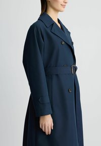 Trench coat blu navy con design doppiopetto, ampio colletto, vita con cinturino e dettagli con bottoni. Tessuto liscio con tasche laterali.