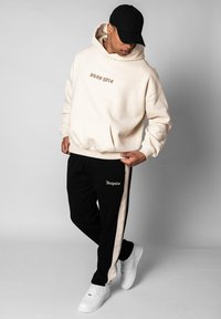 Cremefarbener Kapuzenpullover mit Fronttasche und "Dropsize"-Schriftzug, kombiniert mit schwarzen Jogginghosen, die cremefarbene Seitenstreifen und ein Logo haben. Weiße Sneaker werden getragen.