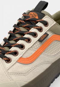 Zapatilla Vans con una parte superior de ante y malla en color beige, acentos en naranja, cordones con patrón negro y naranja, y suela de goma con detalle texturizado.